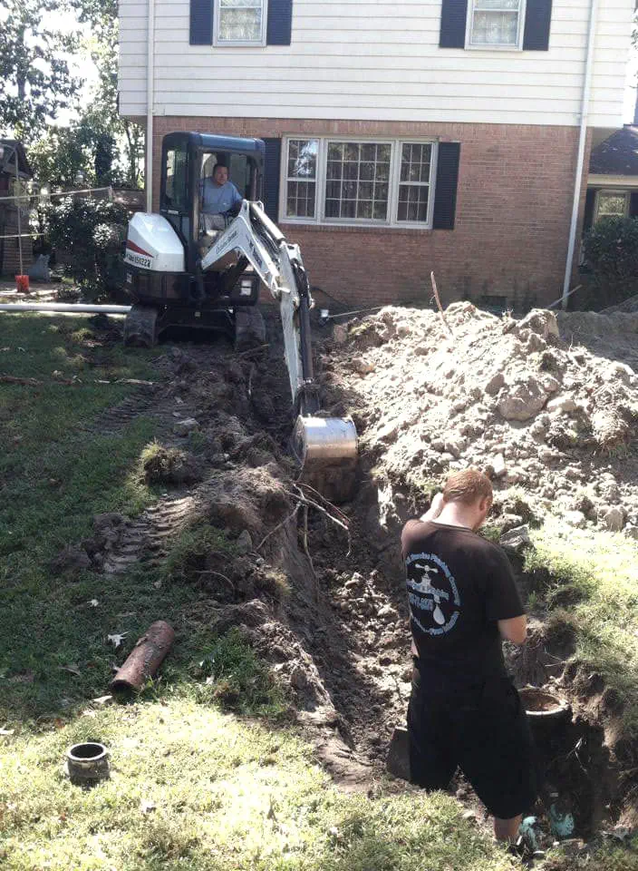 A George's Plumbing tech in a dug trench beside a mini excavator, working on a line at a brick house